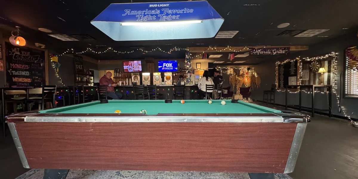 Casual daiquiri bar with pool tables, dart boards and cornhole games visible inside, featuring a busy drive-thru window...