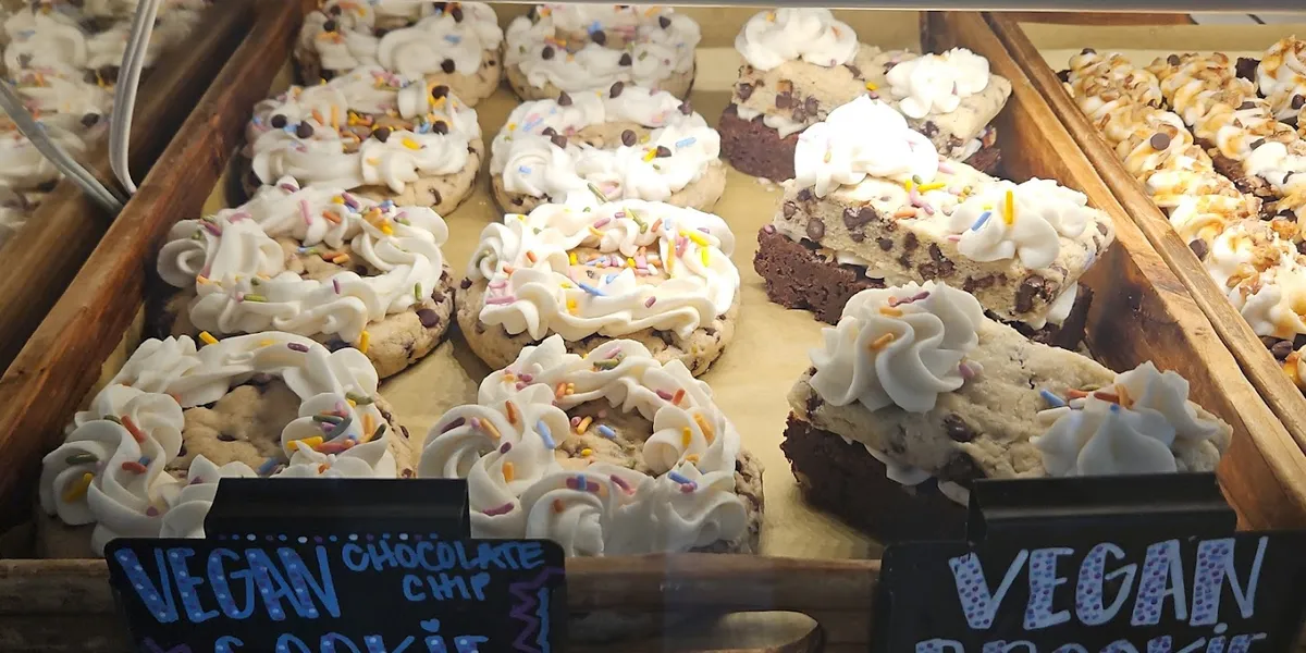 Bright bakery interior with display case showcasing colorful gluten-free and vegan pastries, with outdoor seating visible...