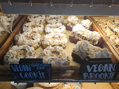 Bright bakery interior with display case showcasing colorful gluten-free and vegan pastries, with outdoor seating visible...
