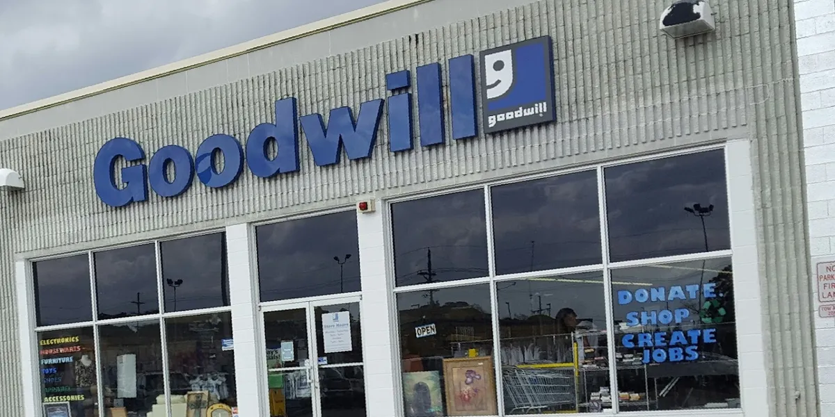 Compact thrift store with organized racks of secondhand clothing, household items, and electronics under fluorescent...