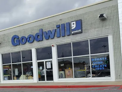 Compact thrift store with organized racks of secondhand clothing, household items, and electronics under fluorescent...