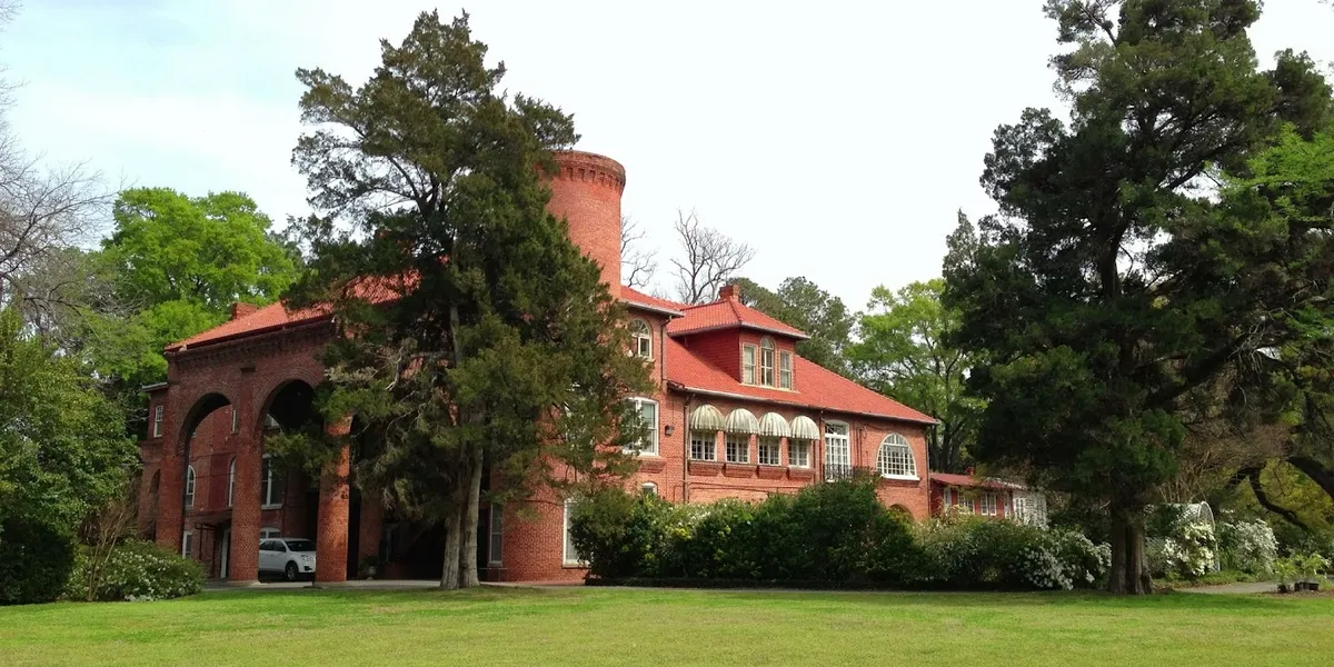 Stately 19th-century southern mansion with sprawling grounds, ancient trees, and ornate architectural details bathed in...