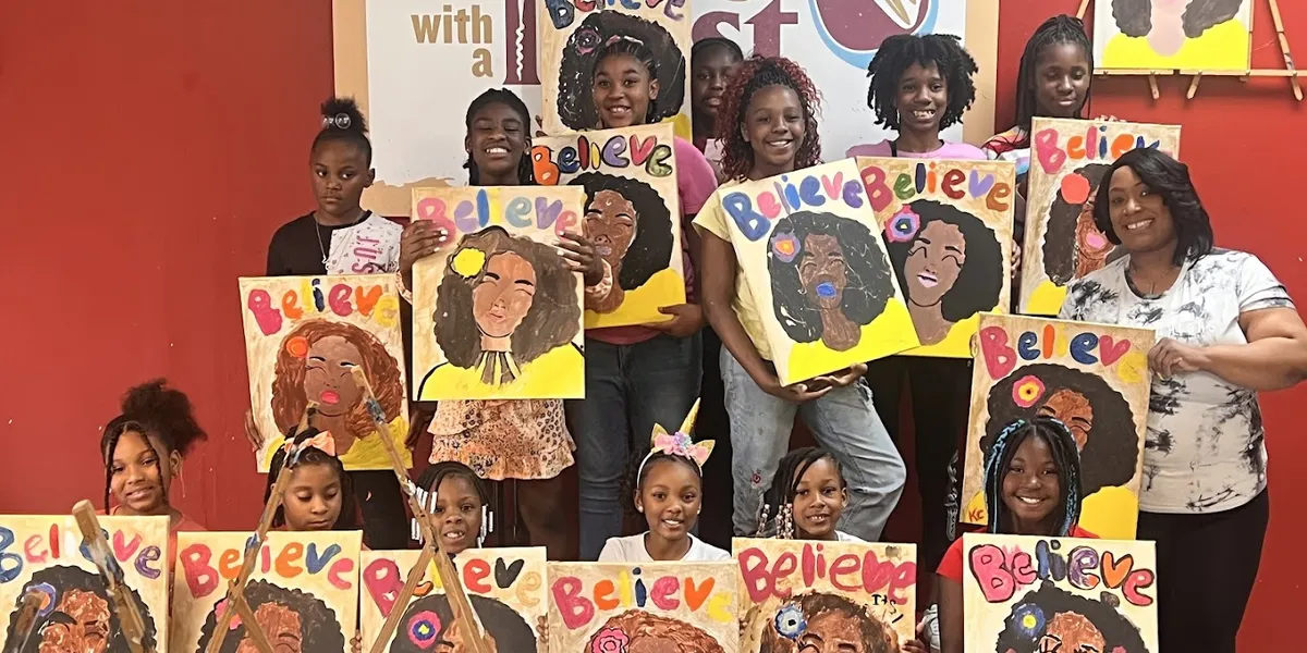 Bright art studio with canvas stations arranged in rows, paint supplies at each seat, and colorful sample paintings...