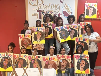 Bright art studio with canvas stations arranged in rows, paint supplies at each seat, and colorful sample paintings...