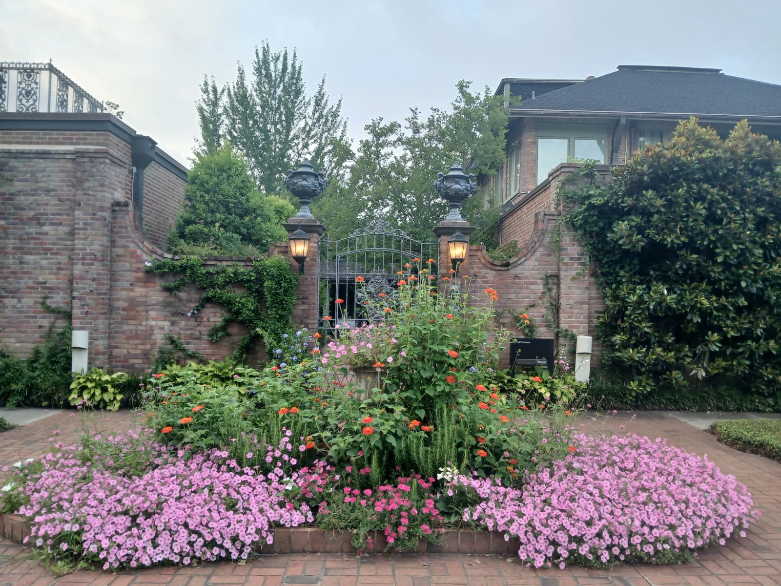Elsong Gardens entrance at Biedenharn Museum Monroe Louisiana with pink petunias orange zinnias and ornate iron gate between brick walls with gas lanterns