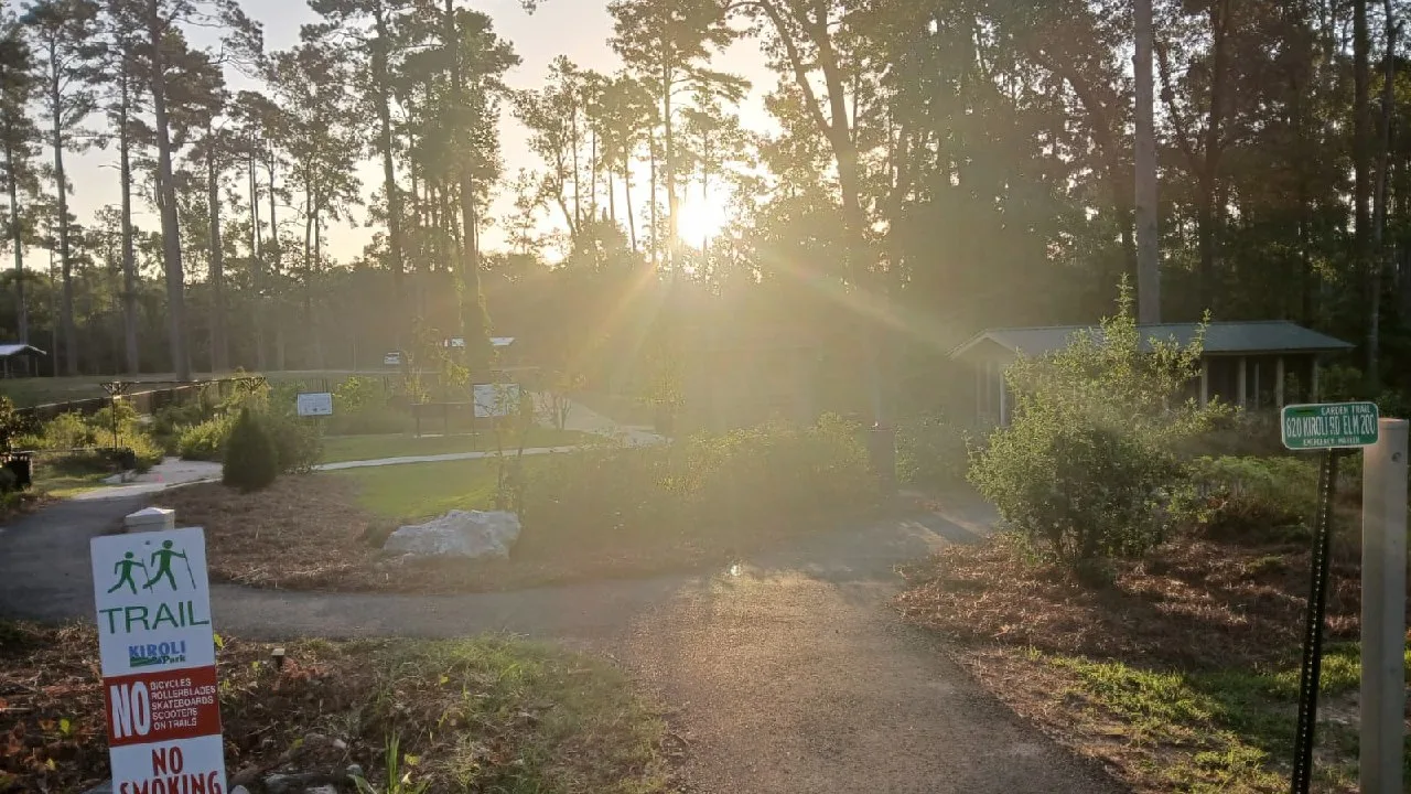 A paved trail lined with park signs winds through tall pine trees at Kiroli Park in West Monroe, Louisiana.