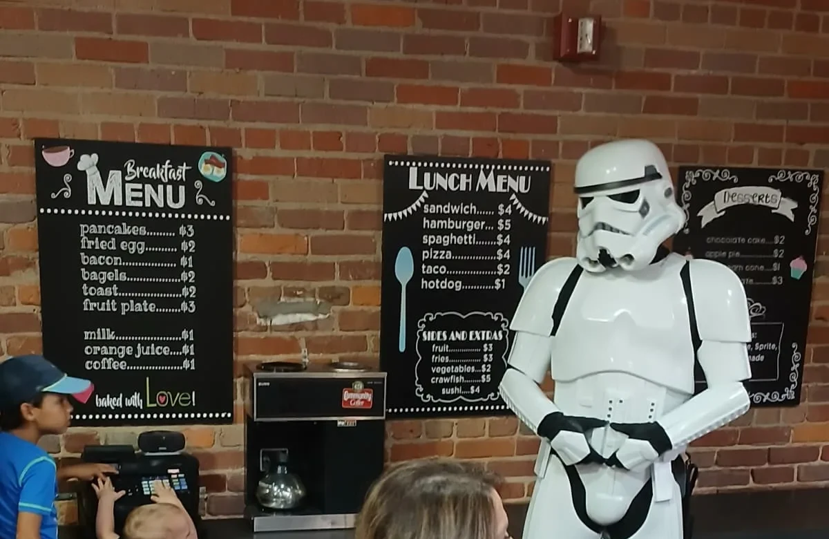 Stormtrooper from the 501st Legion standing in the Kids Cafe exhibit at the Northeast Louisiana Children's Museum, surrounded by chalkboard menus listing pancakes and crawfish